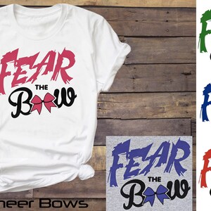 May include: A white t-shirt with the text "FEAR THE BOW" in pink and black. The word "FEAR" is in a bold, stylized font with a pink, brushstroke effect. The word "BOW" is in a black, cursive font with a pink bow in the center. The t-shirt is shown on a wooden background with other t-shirts in different colors with the same design.