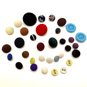 May include: A collection of assorted fabric-covered buttons in various sizes and colors, including black, red, blue, green, brown, white, and yellow. Some buttons feature textured surfaces, patterns, or visible metal eyelets for attachment.