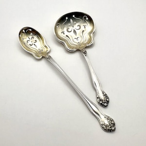May include: Two silver-toned metal serving spoons with ornate, pierced cut-out designs in the bowls. The handles feature detailed floral embossing at the ends, and the bowls have a scalloped edge shape with intricate scrollwork patterns.