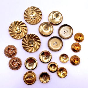 May include: A collection of various gold-toned metal buttons in different sizes and designs. The assortment includes circular buttons with radial sunburst patterns, smooth concave surfaces, raised rims, and intricate braided textures, all featuring two holes.
