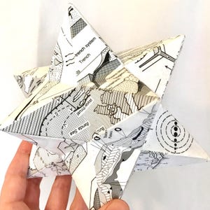 Puede incluir: Una escultura en forma de estrella de papel blanco con un diseño tipo mapa en blanco y negro. La estrella tiene múltiples puntas y está sostenida por una mano. El mapa incluye texto como "Trench system" y "Banda Sea".