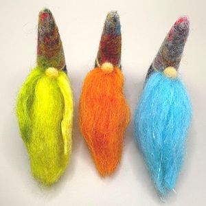 Pode incluir: Três gnomos de feltro com barbas coloridas e chapéus estampados. Os gnomos apresentam barbas amarelas, laranjas e azuis, cada uma com um pequeno nariz bege redondo. Os chapéus são multicoloridos, adicionando ao seu aspecto lúdico.