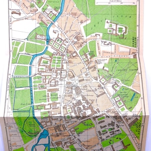 May include: A vintage-style printed map of Cambridge, England, featuring a grid layout with labeled streets, colleges, and green spaces. The map uses a color palette of tan, green, and blue, with a scale of yards and meters in the top right corner.