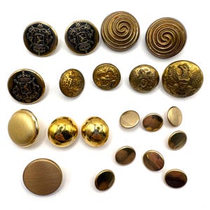 Op de afbeelding: Een verzameling diverse metalen knopen in goud- en messingkleuren. Het assortiment bevat ronde knopen met reliëf heraldische wapens, spiraalpatronen, adelaarmotieven en gladde gepolijste oppervlakken in verschillende maten en afwerkingen.