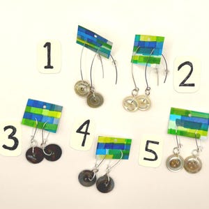 May include: Five pairs of handmade button earrings. Each pair features a different color button, suspended from a silver wire. The earrings are attached to small, colorful, rectangular cards with a blue, green, and yellow design. Numbers 1-5 are on white cards.