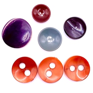 Pode incluir: Uma coleção de botões vintage em várias cores e tamanhos. Os botões incluem tons de roxo, vermelho, laranja e cinza. Alguns botões têm dois orifícios, enquanto outros têm quatro. Os botões estão dispostos sobre um fundo branco.