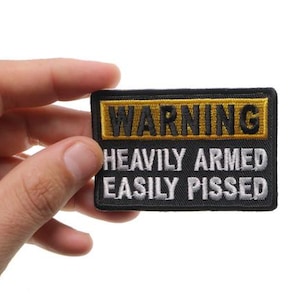 May include: A black and gold embroidered patch with the words "WARNING HEAVILY ARMED EASILY PISSED". The patch is rectangular and has a yellow border around the top.