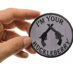 May include: A round, gray embroidered patch with black stitching. The patch features two crossed pistols and the text "I'm Your Huckleberry".
