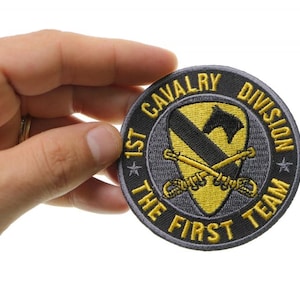 May include: A gray embroidered patch with yellow stitching. The patch features a black horse head inside a yellow diamond shape with crossed swords. The text around the patch reads "1st Cavalry Division The First Team".