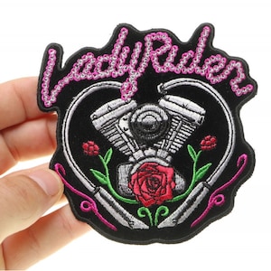 May include: Black embroidered patch with a pink "Lady Rider" text, a stylized heart with a motorcycle engine inside, and a red rose.