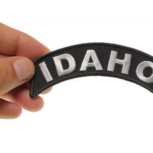 May include: Black and white embroidered patch with the word "IDAHO" in white lettering on a black background.