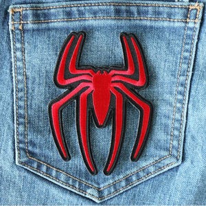 May include: Red and black spider-shaped patch on a blue denim background. The patch is sewn onto the back pocket of a pair of jeans.
