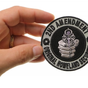 May include: A black and white embroidered patch with the text "2nd Amendment Original Howland Security" and a stylized image of a hand holding a gun.