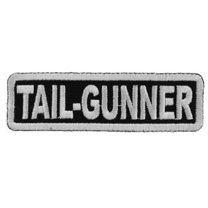 May include: A rectangular black patch with white embroidered text that reads "TAIL-GUNNER". The text is in a bold, sans-serif font and is outlined with a white border. The patch is likely designed to be sewn or ironed onto clothing or gear.