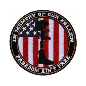 May include: A round black and gold embroidered patch with an American flag in the center. The flag has red and white stripes and white stars on a blue field. A silhouette of a soldier in a helmet is standing in front of the flag, holding a rifle. The text "In Memory of Our Fallen" is written in a circle around the top of the patch, and the text "Freedom Ain't Free" is written in a circle around the bottom of the patch.