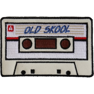 May include: White cassette tape with black trim and the text "OLD SKOOL" in blue lettering. The cassette tape has a red and white stripe across the top and a brown rewind button on each side.