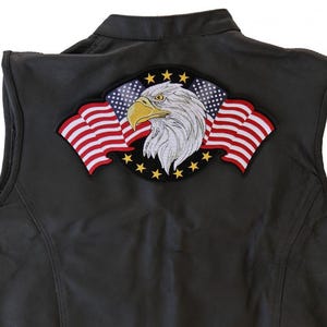 May include: Black leather vest featuring a large embroidered patch of an American bald eagle with the American flag. The patch is centered on the back of the vest. The vest has a collar and a sleeveless design.