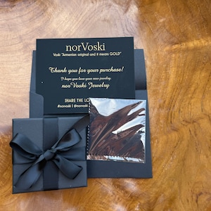 May include: A black gift box with a black ribbon tied in a bow. The box is sitting on a wooden surface. A black card with gold lettering reads "nor Voski Voski "Armenian original and it means GOLD" Thank you for your purchase! I hope you love your new jewelry nor Voski Jewelry SHARE THE LOVE #norvoski @norvoski"