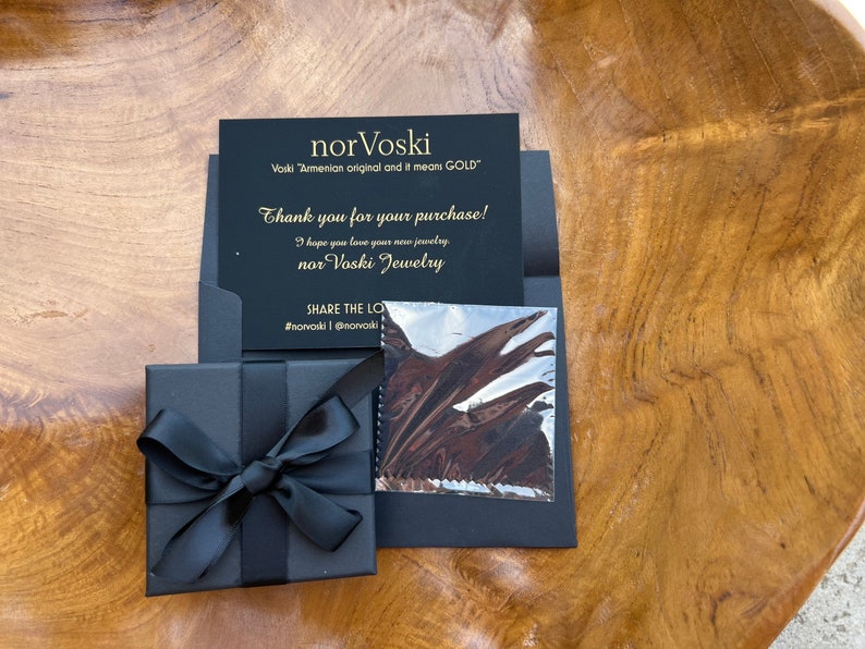 May include: A black gift box with a black ribbon tied in a bow. The box is sitting on a wooden surface. A black card with gold lettering reads "nor Voski Voski "Armenian original and it means GOLD" Thank you for your purchase! I hope you love your new jewelry nor Voski Jewelry SHARE THE LOVE #norvoski @norvoski"