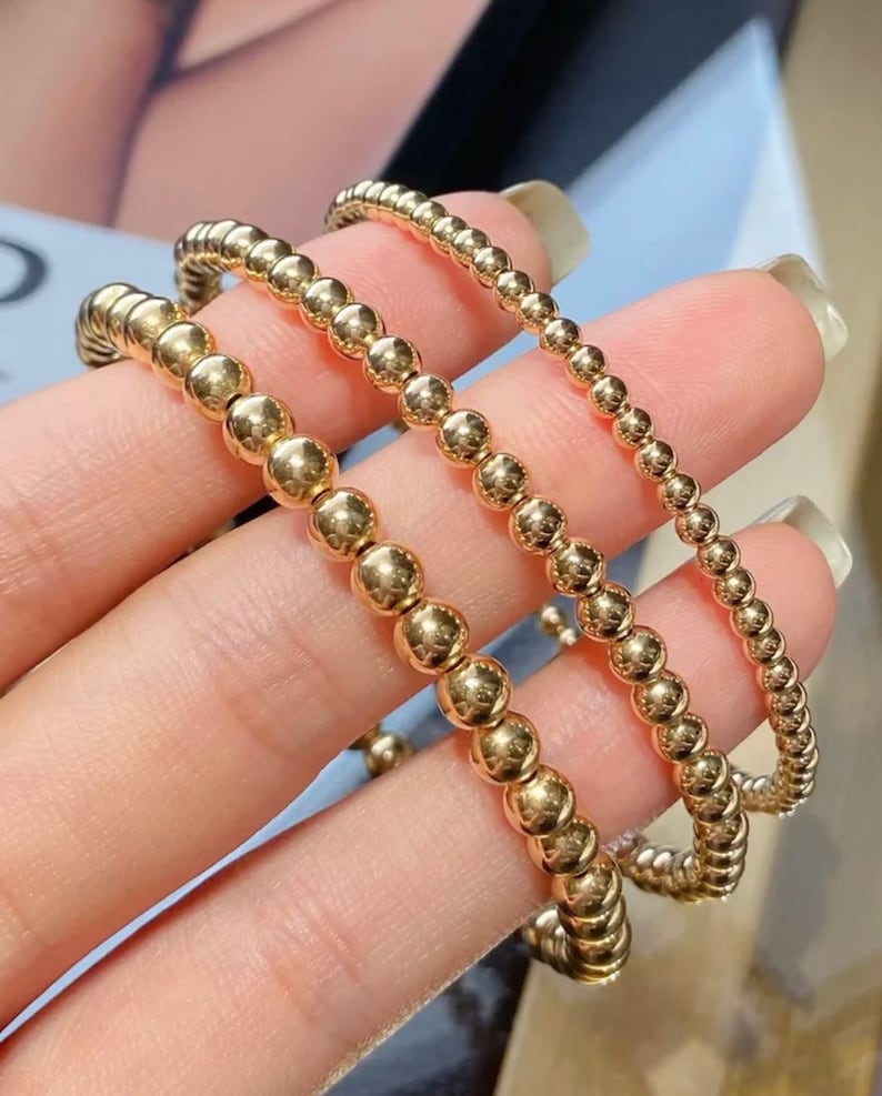 May include: Three gold beaded bracelets of varying sizes stacked on top of each other.
