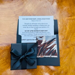 May include: A black gift box with a black ribbon tied in a bow. The box is sitting on a wooden surface. A card with the text "YOU ARE VOSKI BABY, A REAL SOLID VOSKI Enjoy it, live in it. Though you will never worry about your norVoski Jewelry tarnishing or fading here are some tips on how to keep your jewelry at its finest PAT DRY AFTER SHOWER TO KEEP SHINY Avoid leaving jewelry on while using abrasive scrubs, exfoliant or cleansers. Though your piece is Solid Gold, often times gold can ty chem ALWAYS supervise kids wha" is next to the box.