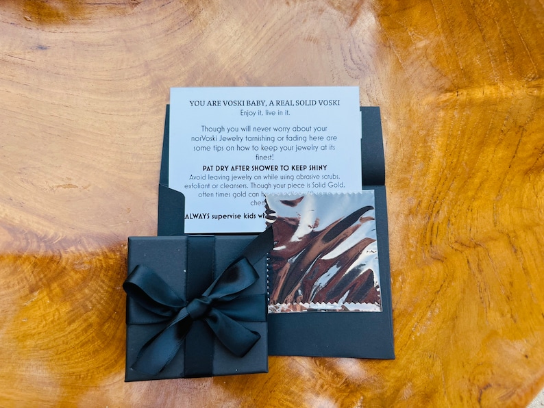 May include: A black gift box with a black ribbon tied in a bow. The box is sitting on a wooden surface. A card with the text "YOU ARE VOSKI BABY, A REAL SOLID VOSKI Enjoy it, live in it. Though you will never worry about your norVoski Jewelry tarnishing or fading here are some tips on how to keep your jewelry at its finest PAT DRY AFTER SHOWER TO KEEP SHINY Avoid leaving jewelry on while using abrasive scrubs, exfoliant or cleansers. Though your piece is Solid Gold, often times gold can ty chem ALWAYS supervise kids wha" is next to the box.
