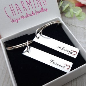 May include: A silver keyring with two rectangular tags. The tags are engraved with the words "Forever" and "Always" with a small heart symbol next to each word. The keyring is in a white box with the words "How Charming Unique Handmade Jewellery" printed on the lid.