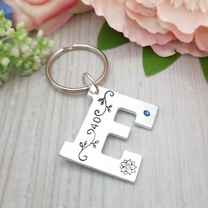 Könnte beinhalten: Silberner Schlüsselanhänger mit einem Anhänger in Form des Buchstabens E. Der Anhänger hat ein florales Design und einen kleinen blauen Strassstein. Der Schlüsselanhänger ist an einem silbernen Schlüsselring befestigt.