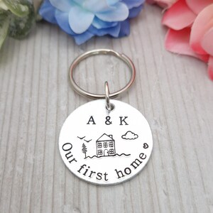 以下が含まれることがあります： 家のデザインが刻まれたシルバーのキーホルダー。"A & K Our first home"の文字が刻まれています。