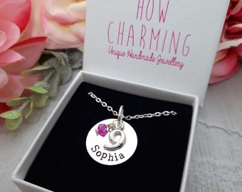 Op de afbeelding: Een zilveren ketting met een ronde hanger waarop "Sophia" staat en het nummer "9". De hanger heeft een roze kristal. De ketting zit in een witte doos met de tekst "HOW CHARMING Unique Handmade Jewellery" erop.
