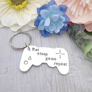 May include: Silver-colored keychain in the shape of a video game controller. The keychain has the words "Eat sleep game repeat" stamped on it. The keychain is next to white, blue, and pink flowers.