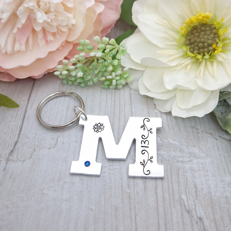 May include: Silver metal keychain featuring the letter "M". The letter has a floral design, a blue gem, and the numbers "913C". The keychain is attached to a silver ring. The background includes white and pink flowers.
