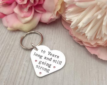 May include: A silver-coloured heart-shaped metal keyring with a split ring. The surface is engraved with the black text 10 Years long and still going strong, accented by three small red heart symbols stamped below the text.