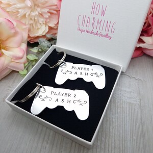 Op de afbeelding: Twee zilveren sleutelhangers in de vorm van videogamecontrollers. De sleutelhangers bevinden zich in een witte doos met een zwarte voering. De tekst op de sleutelhangers luidt "Player 1 A & H" en "Player 2 A & H".