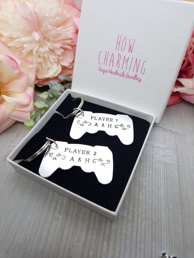 Op de afbeelding: Twee zilveren sleutelhangers in de vorm van videogamecontrollers. De sleutelhangers bevinden zich in een witte doos met een zwarte voering. De tekst op de sleutelhangers luidt "Player 1 A & H" en "Player 2 A & H".