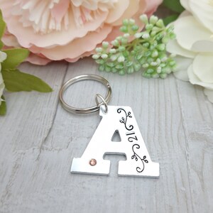May include: Silver metal keychain in the shape of the letter "A". The letter has a small orange gem and black floral design. The number "21" is also on the keychain. The keychain is attached to a silver metal ring.