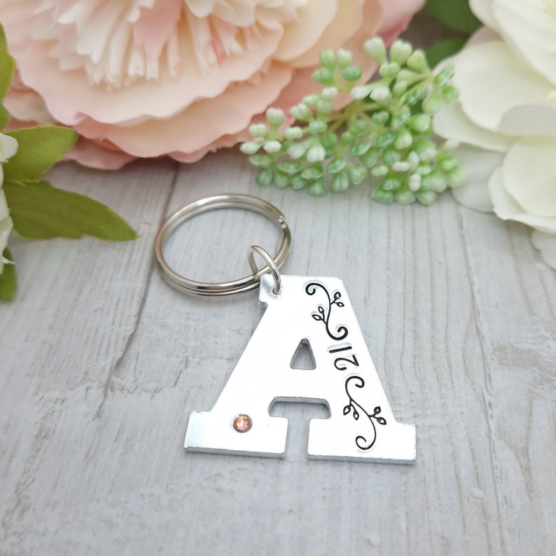 May include: Silver metal keychain in the shape of the letter "A". The letter has a small orange gem and black floral design. The number "21" is also on the keychain. The keychain is attached to a silver metal ring.