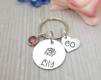 May include: A silver keychain with a round charm engraved with a rose and the name "Lily", a heart charm with the number "60", and a pink gemstone. The keychain is on a silver ring.