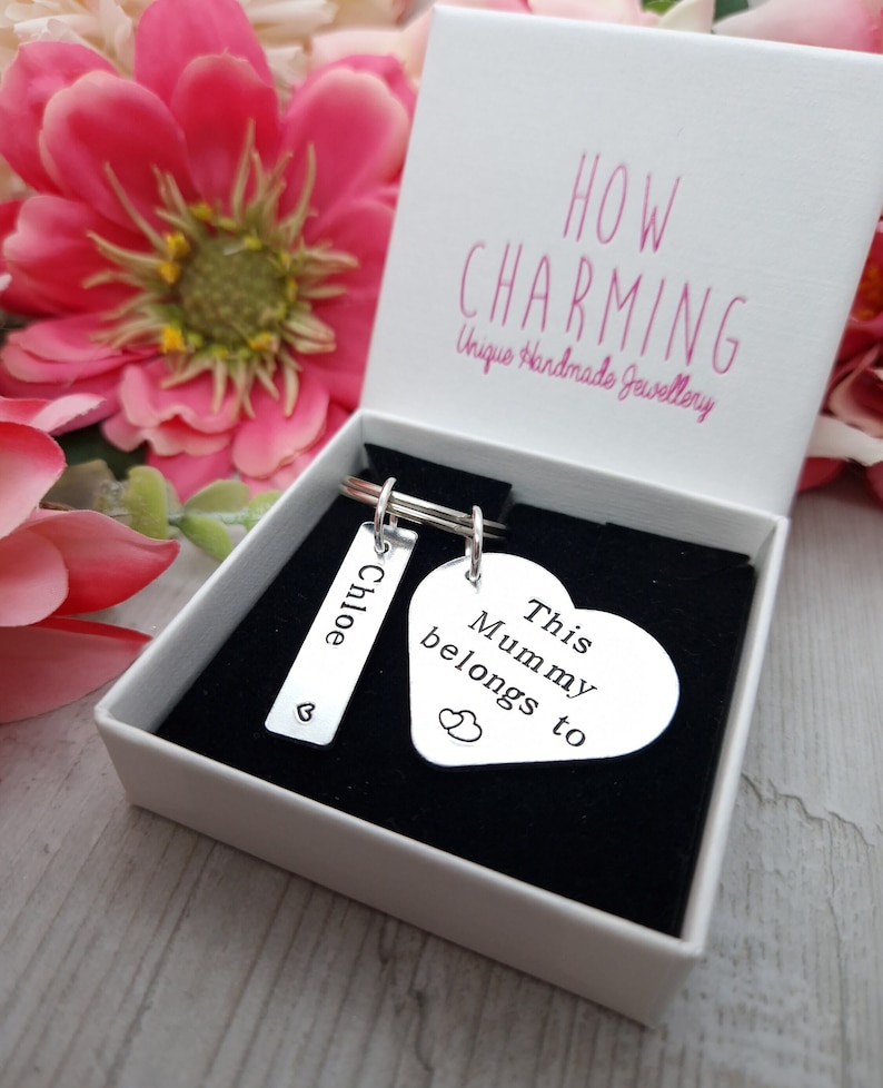 May include: A silver heart-shaped keyring with the text "This Mummy belongs to" and two small hearts. A silver rectangular tag with the name "Chloe" and a small heart is attached to the keyring. The keyring is in a white box with the text "HOW CHARMING Unique Handmade Jewellery".