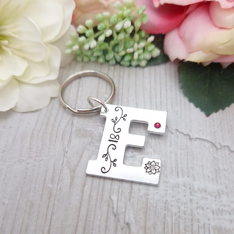 May include: Silver metal keychain in the shape of the letter "E". The letter is decorated with black floral designs and a small red gem. A small flower design is also present. The keychain ring is silver.