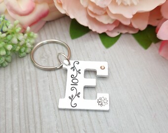 May include: Silver keyring with a letter E charm. The charm has a floral design and a small rhinestone. The keyring is attached to a silver key ring.