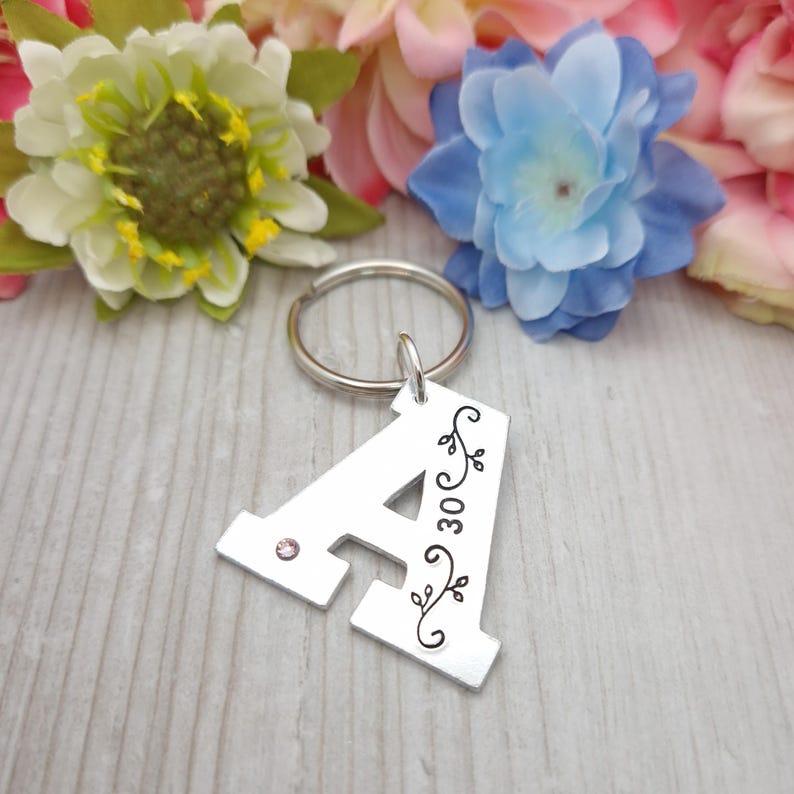 May include: A silver metal keychain in the shape of the letter "A". The letter has a small pink gem and black floral design with the number "30". The keychain is attached to a silver ring. Colorful flowers are in the background.