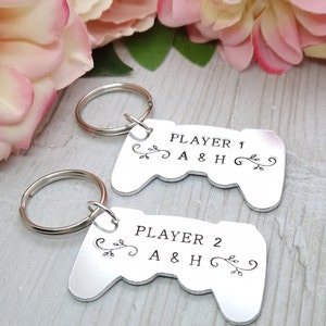 Op de afbeelding: Twee zilveren sleutelhangers in de vorm van videogamecontrollers. Elke sleutelhanger heeft een zwarte inscriptie met "Player 1 A & H" en "Player 2 A & H".