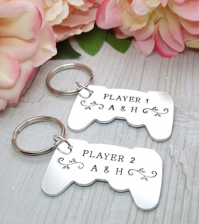 Op de afbeelding: Twee zilveren sleutelhangers in de vorm van videogamecontrollers. Elke sleutelhanger heeft een zwarte inscriptie met "Player 1 A & H" en "Player 2 A & H".