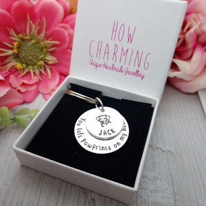 May include: A silver keyring with a round charm that says "You left paw prints on my heart" and has a small dog silhouette with the name "Jack" on it. The keyring is in a white box with the words "How Charming Unique Handmade Jewellery" printed on it.