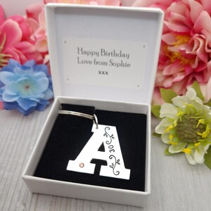 May include: A silver letter "A" keychain with a small pink gem and decorative floral accents, presented in a white gift box. The box contains a message that reads "Happy Birthday Love from Sophie". The background features colorful flowers.