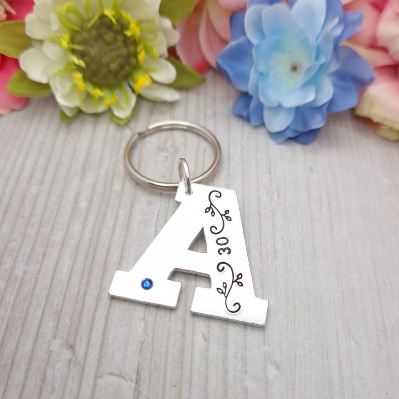 May include: Silver metal keychain in the shape of the letter "A". The letter has a blue gem and a black floral design with the number "30". The keychain is attached to a silver ring.