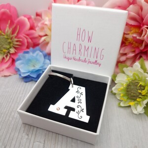 May include: A silver letter "A" keychain with a decorative floral design and the number "30" is displayed in an open white gift box with a black interior. The box has the text "HOW CHARMING Unique Handmade Jewellery" printed on the inside of the lid.