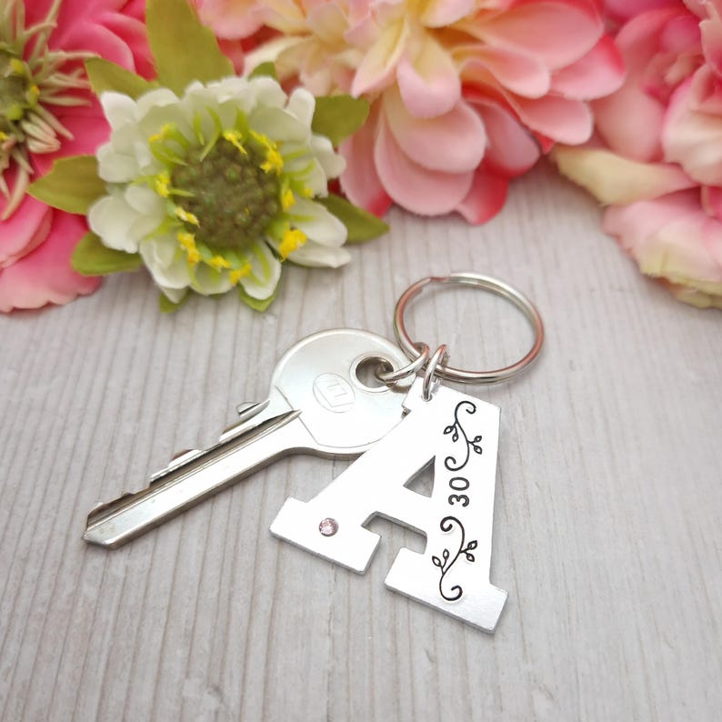 May include: A silver metal keychain with the letter "A" and the number "30" engraved on it. The keychain has a small pink gem and decorative floral accents. A silver key is attached to the keychain. Pink and white flowers are in the background.
