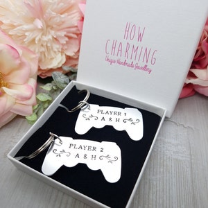 Op de afbeelding: Twee zilveren sleutelhangers in de vorm van videogamecontrollers met de tekst "PLAYER 1 A&H" en "PLAYER 2 A&H" erop. De sleutelhangers zitten in een witte doos met een zwarte binnenkant.